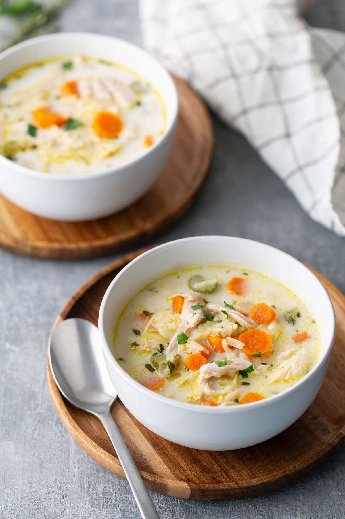 Two bowls of creamy chicken soup with carrots and herbs on wooden trays. A spoon is placed next to one bowl, and a checkered cloth is in the background.