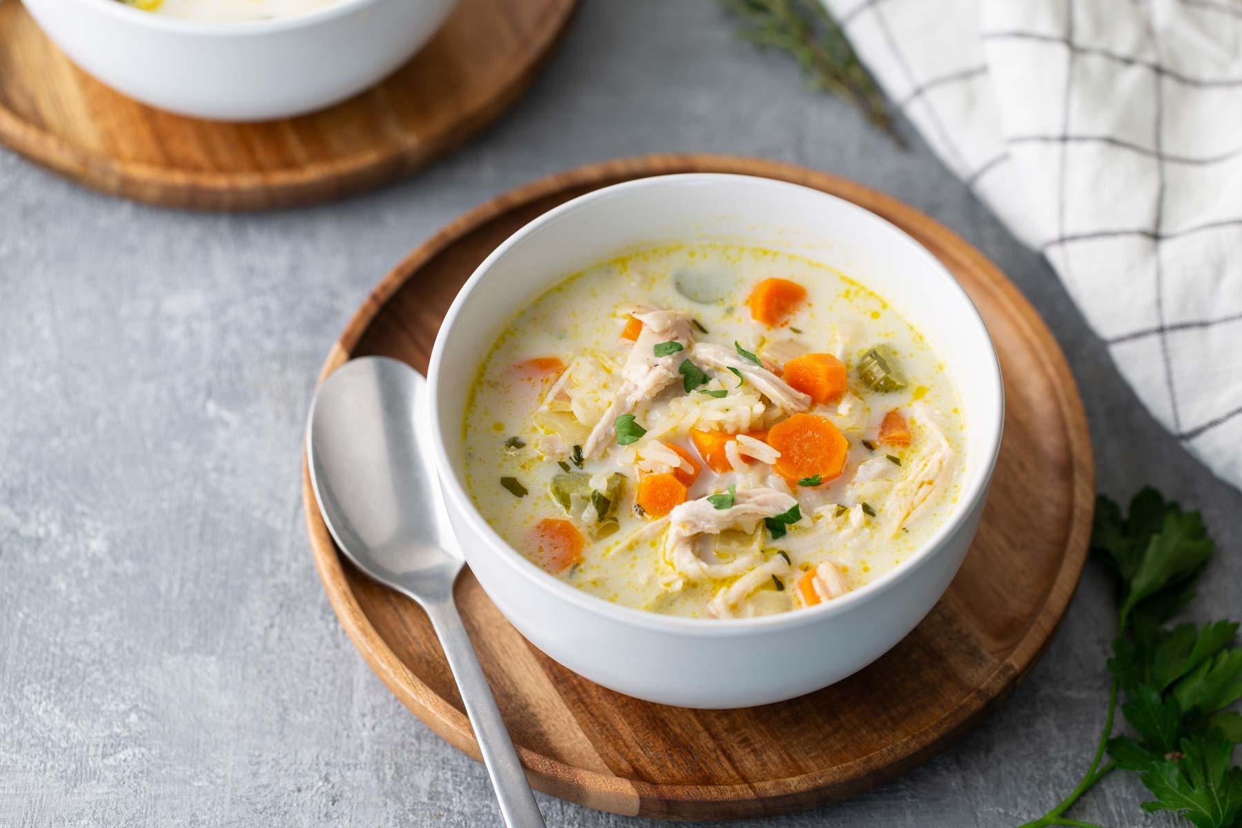 A bowl of creamy chicken noodle soup with carrots, celery, and herbs on a wooden plate with a spoon beside it.