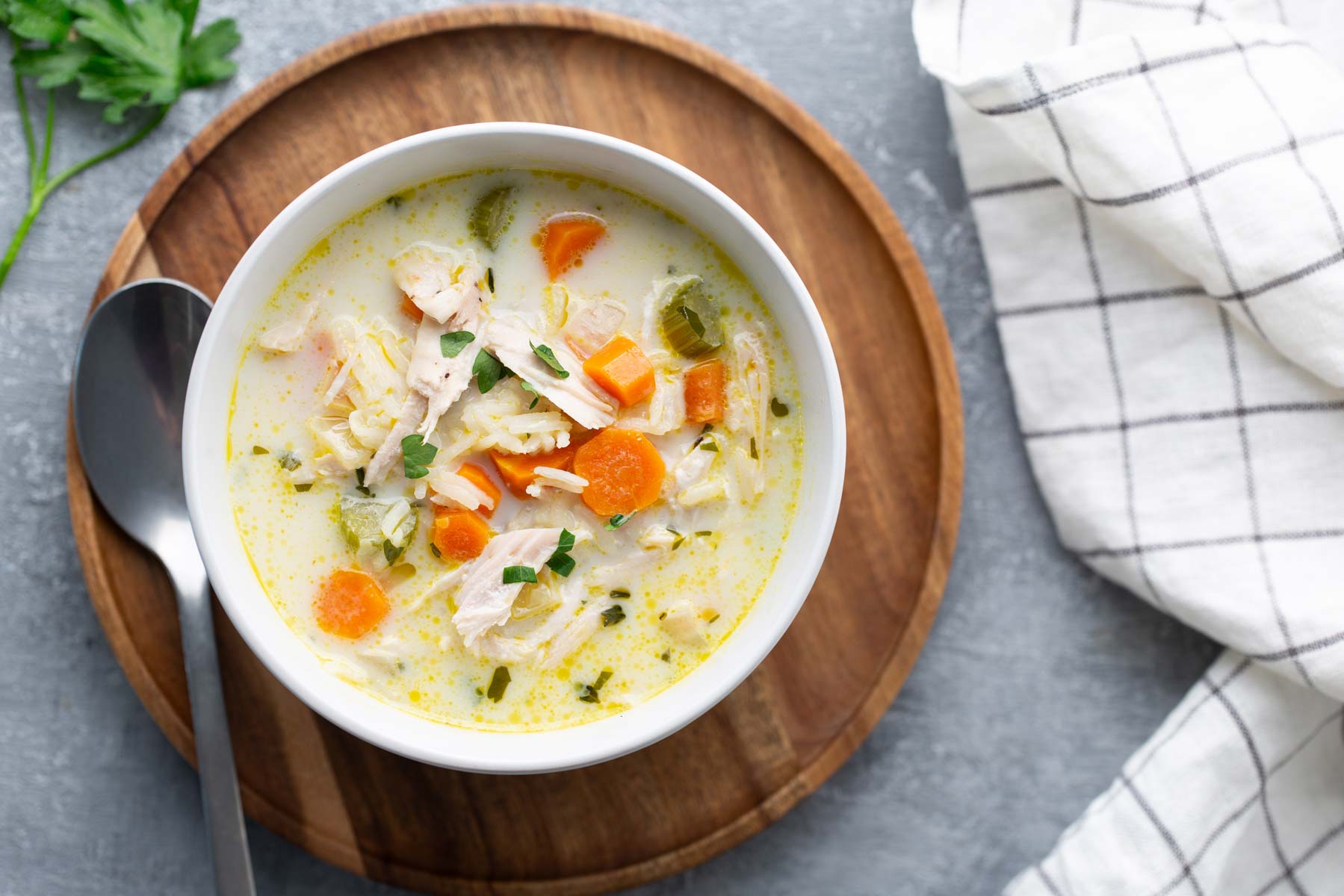 A bowl of creamy chicken soup with carrots, celery, and herbs on a wooden tray, accompanied by a spoon and a checked cloth.