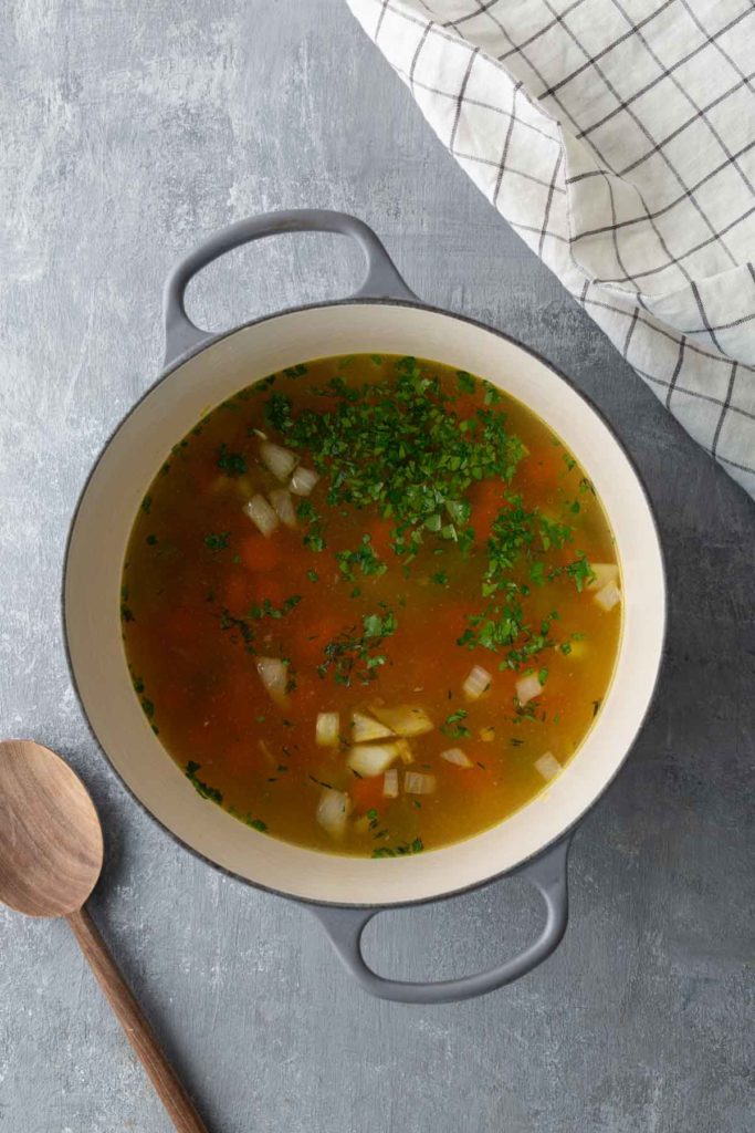 A pot of clear soup with diced onions and chopped parsley on a grey surface, next to a wooden spoon and a checkered cloth.