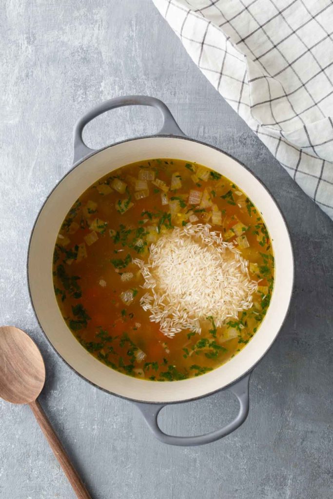 A pot of soup with chunks of vegetables and rice being cooked, next to a wooden spoon and a checkered cloth.