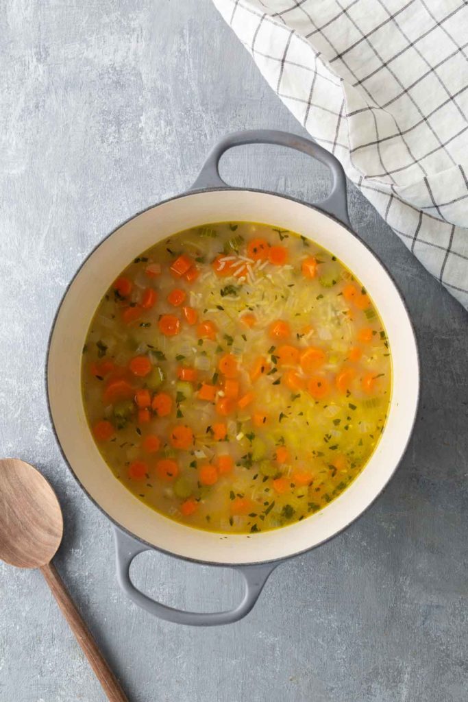 A pot of vegetable soup with carrots, rice, and herbs on a gray surface next to a wooden spoon and a checked cloth.