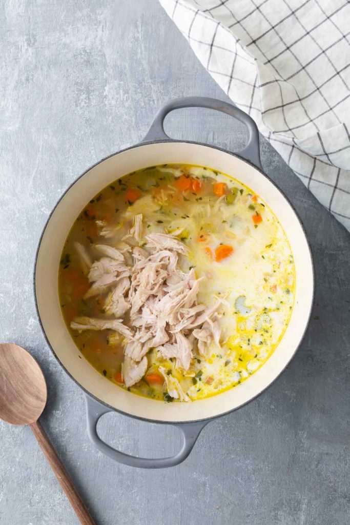 A pot of creamy chicken and vegetable soup with shredded chicken on top. A wooden spoon and a checkered cloth are beside the pot.