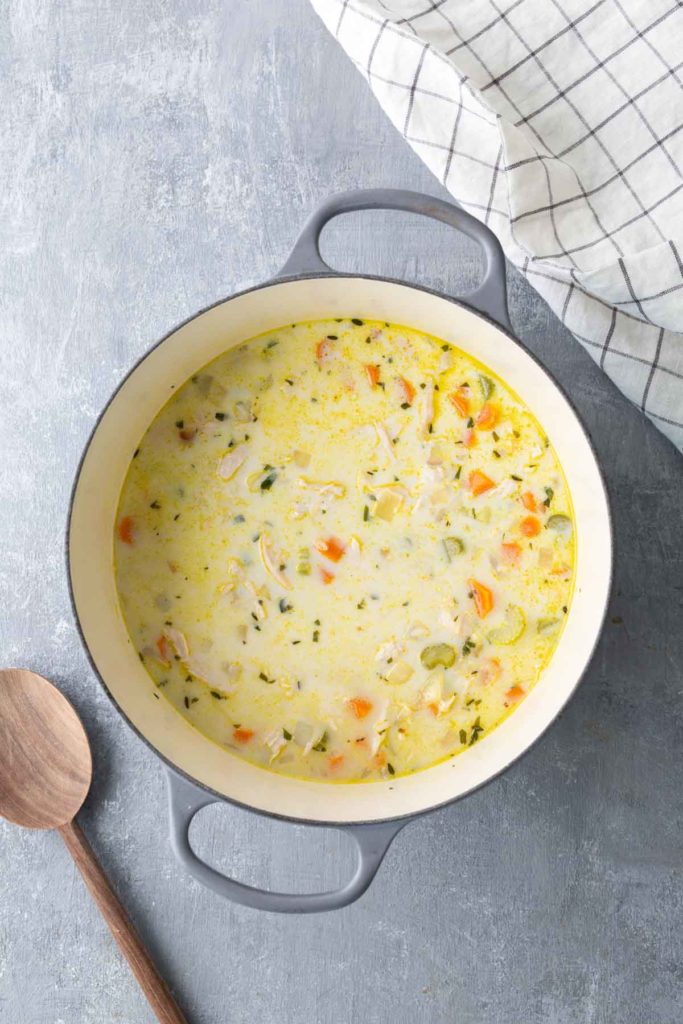 A pot of creamy soup with vegetables and chicken, next to a wooden spoon and a checkered cloth on a gray surface.