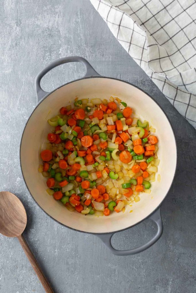 A pot with chopped carrots, celery, and onions on a gray surface next to a wooden spoon and a checkered cloth.