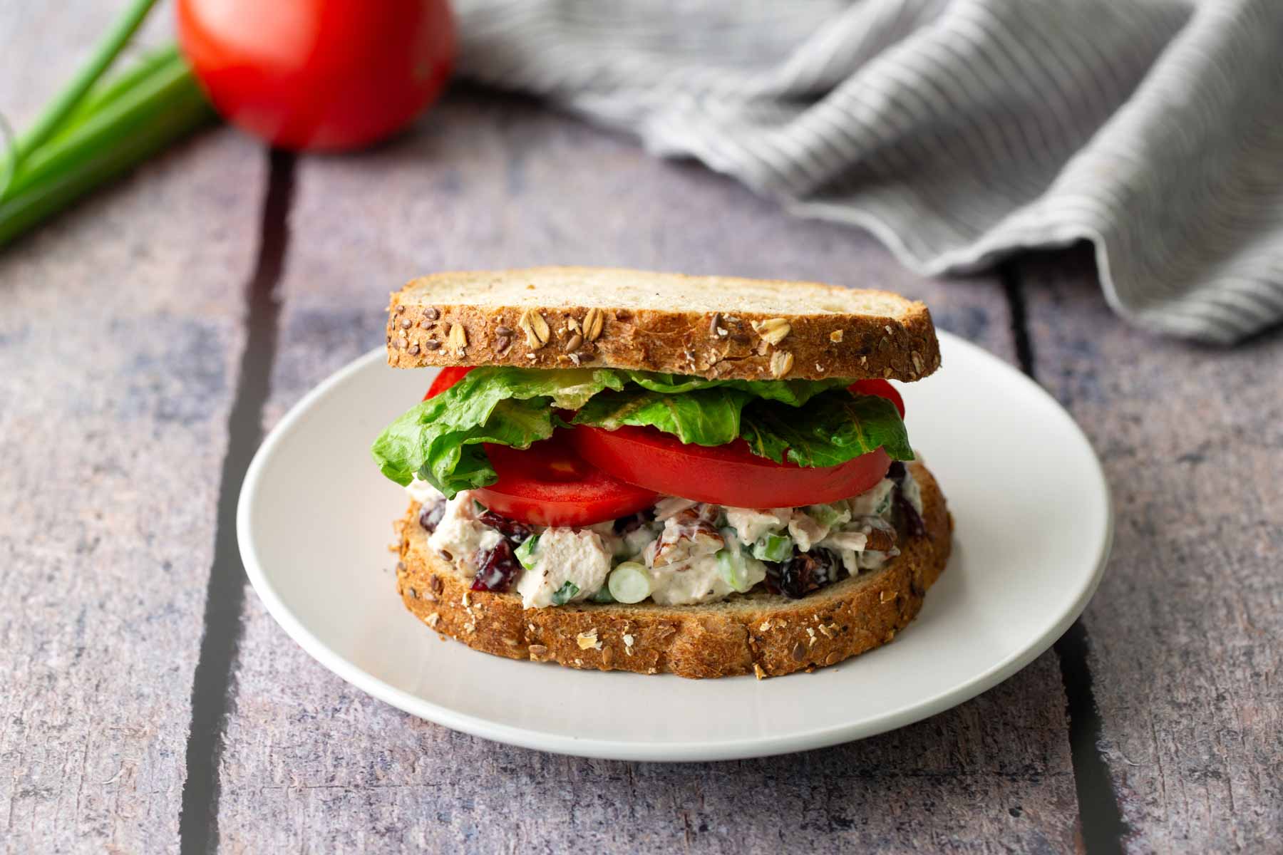 A chicken salad sandwich with lettuce and tomato on whole grain bread sits on a white plate. A tomato and striped cloth are in the background.