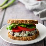 A chicken salad sandwich with lettuce and tomato on whole grain bread sits on a white plate. Background items include a tomato, green onions, and a striped cloth.