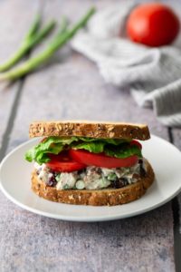 A chicken salad sandwich with lettuce and tomato on whole grain bread sits on a white plate. Background items include a tomato, green onions, and a striped cloth.