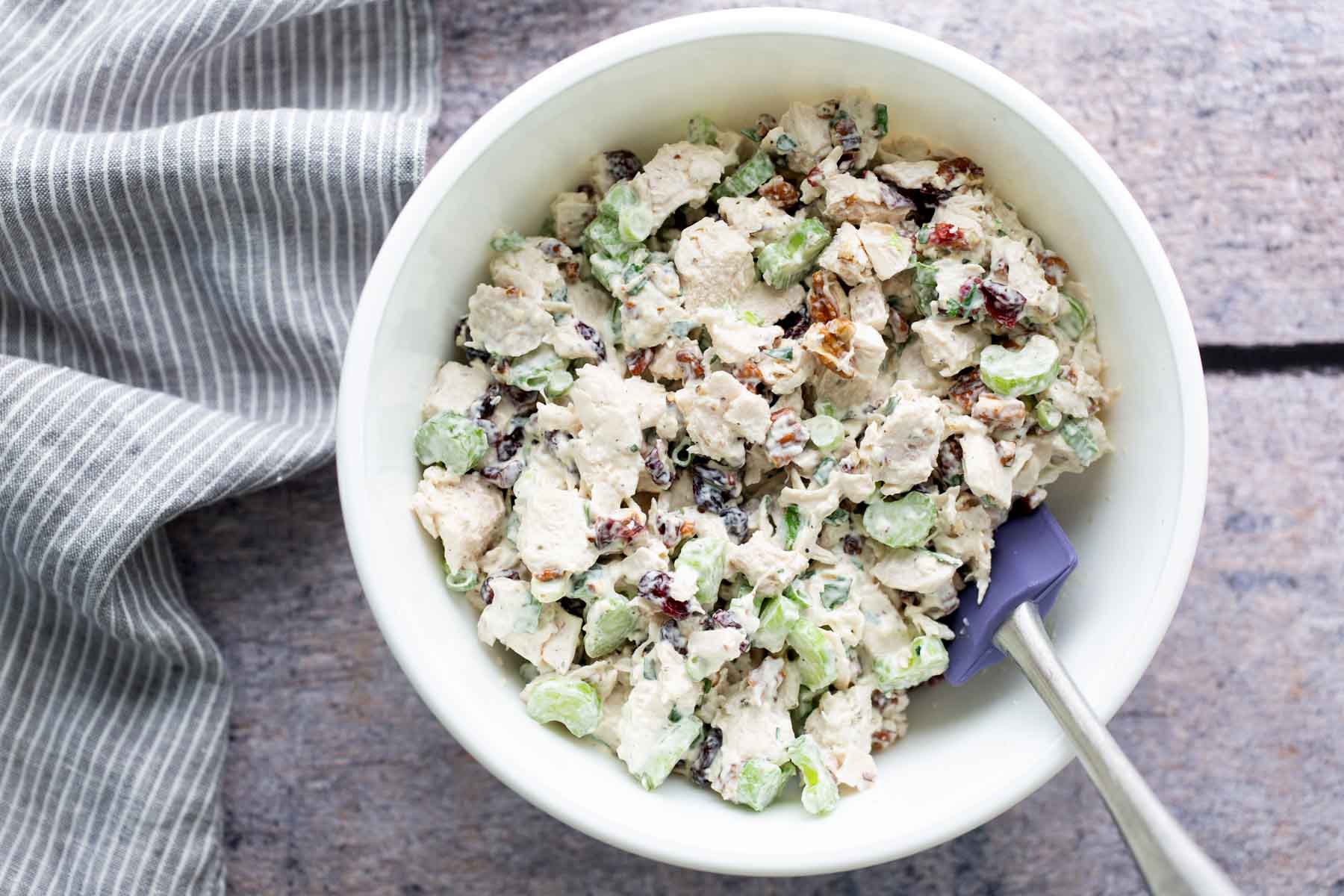 A white bowl contains chicken salad with grapes, nuts, and celery. A striped cloth is next to the bowl.