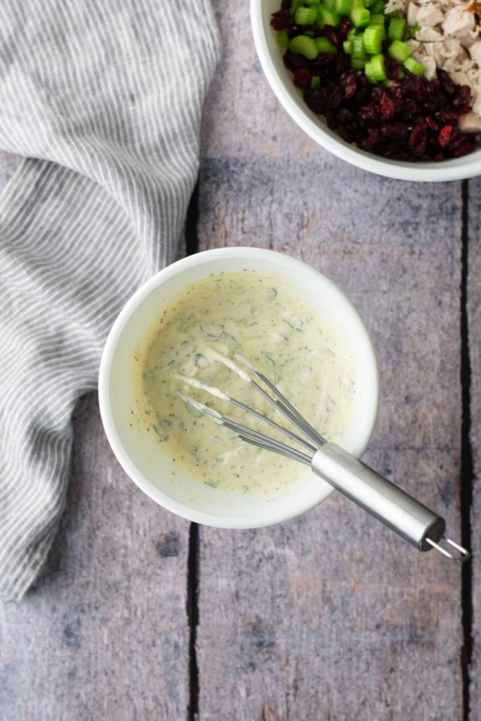 A bowl of creamy dressing with a whisk on a wooden surface. Another bowl with chopped vegetables and dried fruits is partially visible. A striped cloth is placed nearby.