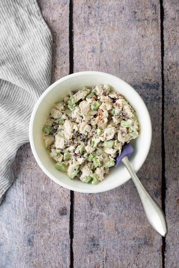 A bowl of chicken salad with sliced celery and herbs, placed on a wooden surface next to a striped cloth.