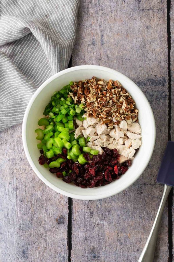 Bowl with diced celery, green onions, chopped pecans, cooked chicken pieces, and dried cranberries on a wooden surface. Striped cloth and spatula nearby.