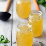 Three glass jars filled with yellow broth are on a wooden surface, surrounded by herbs and a carrot in the background.