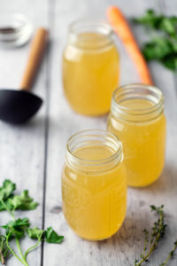 Three glass jars filled with yellow broth are on a wooden surface, surrounded by herbs and a carrot in the background.