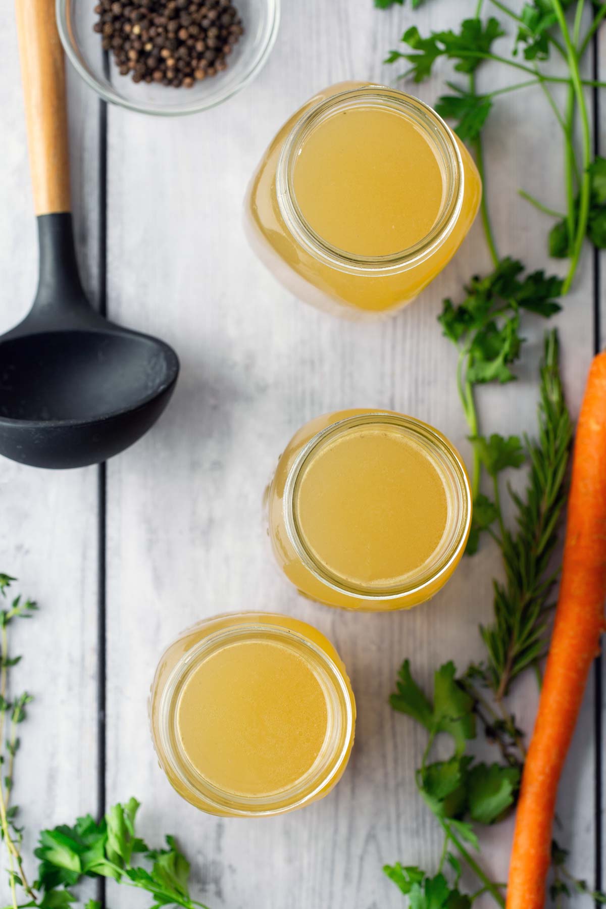 Top view of three jars of broth on a wooden surface, surrounded by a ladle, peppercorns, parsley, a carrot, and rosemary.