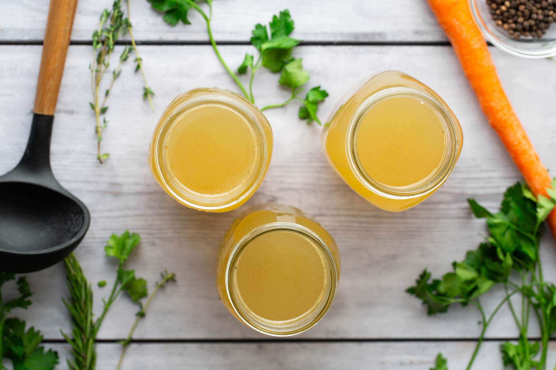 Three jars filled with light golden liquid on a wooden table, surrounded by herbs, carrots, and spices, with a black ladle on the side.