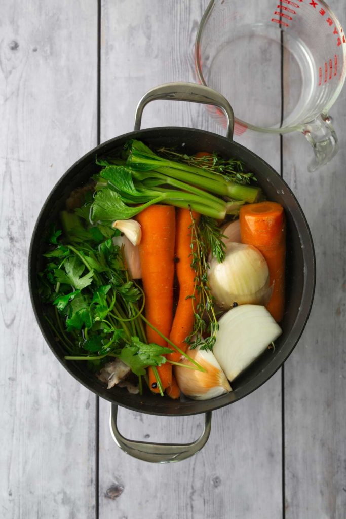 Pot with carrots, onions, celery, parsley, garlic, and herbs on a wooden surface. A clear measuring cup with water is nearby.