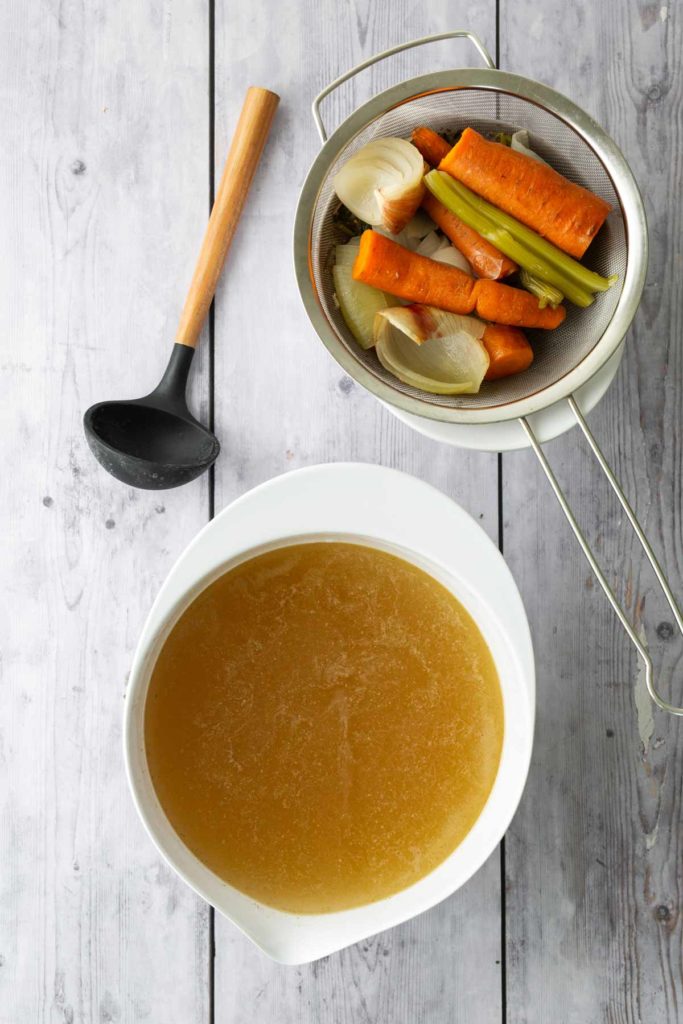 A bowl of broth on a wooden surface with a strainer containing cooked carrots, onions, and celery alongside a black ladle.