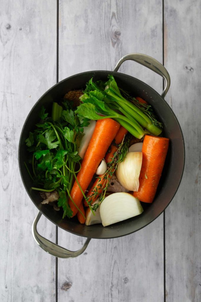 A pot filled with raw ingredients, including carrots, onions, celery, and herbs, over a wooden surface.