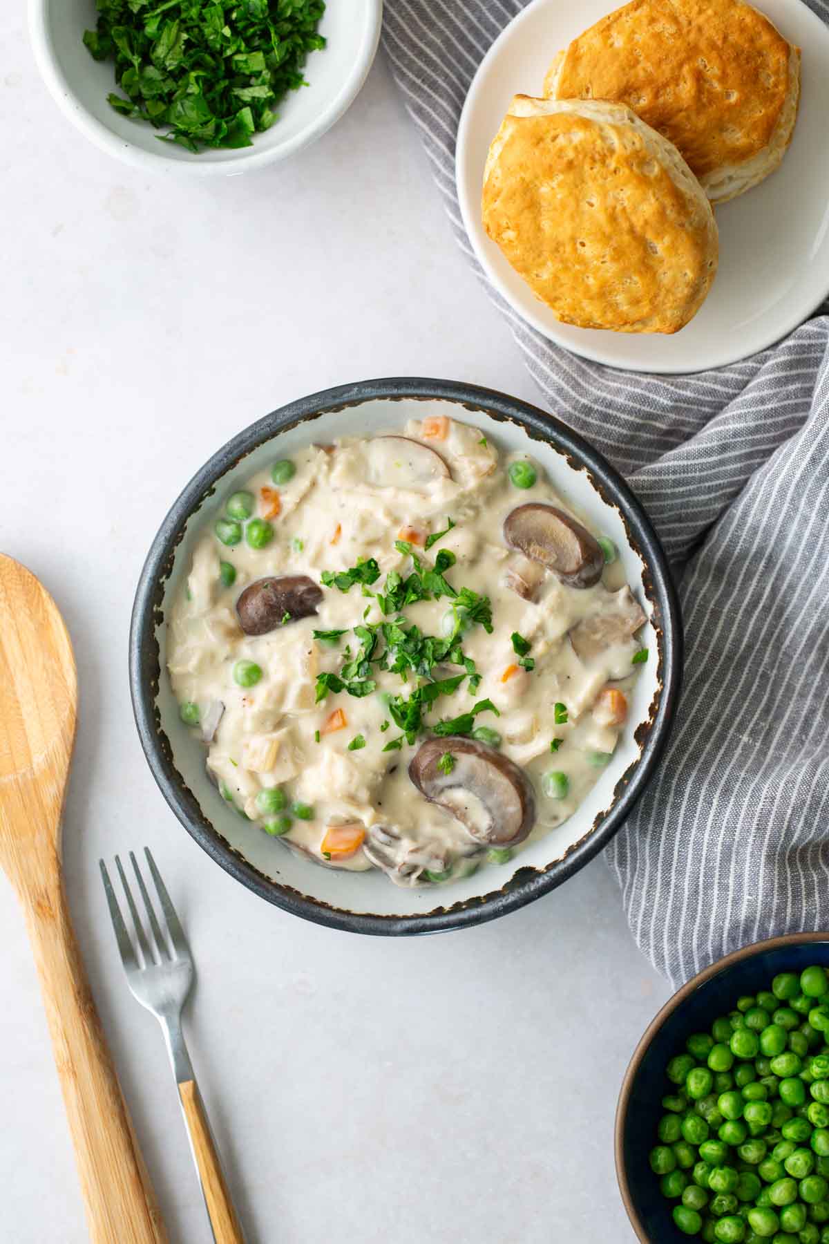 Bowl of creamy chicken and vegetable stew, topped with parsley, next to a striped cloth. Fluffy biscuits and chopped parsley in separate bowls. Wooden spoon and fork on the side.