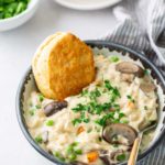 A bowl of creamy chicken and vegetable stew topped with parsley, accompanied by a biscuit. A striped napkin and a plate with more biscuits are in the background.