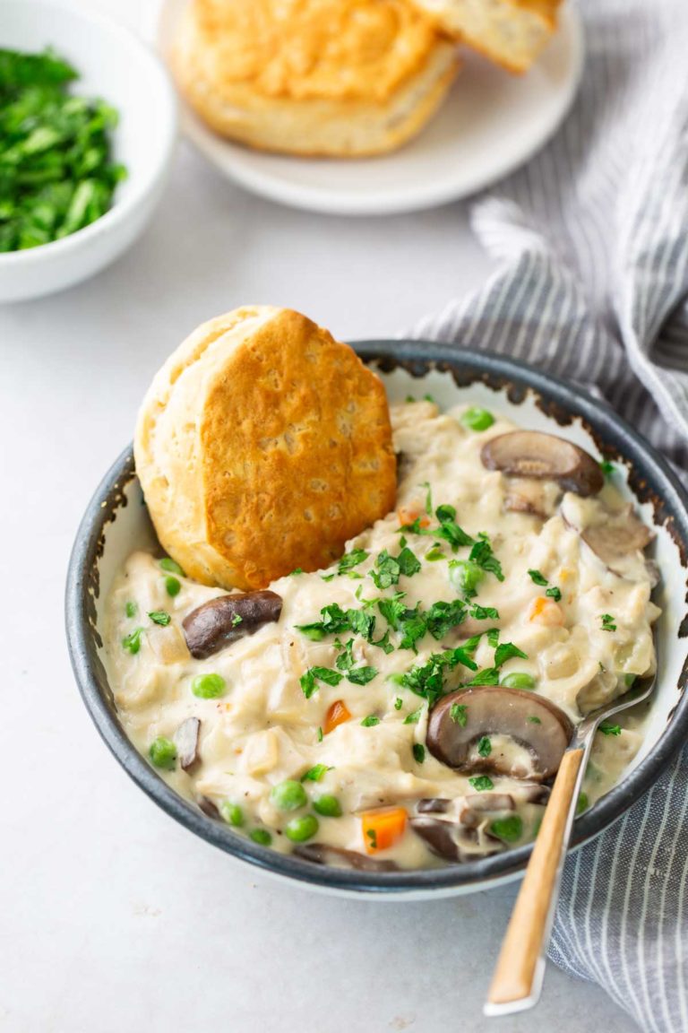 A bowl of creamy chicken and vegetable stew topped with parsley, accompanied by a biscuit. A striped napkin and a plate with more biscuits are in the background.