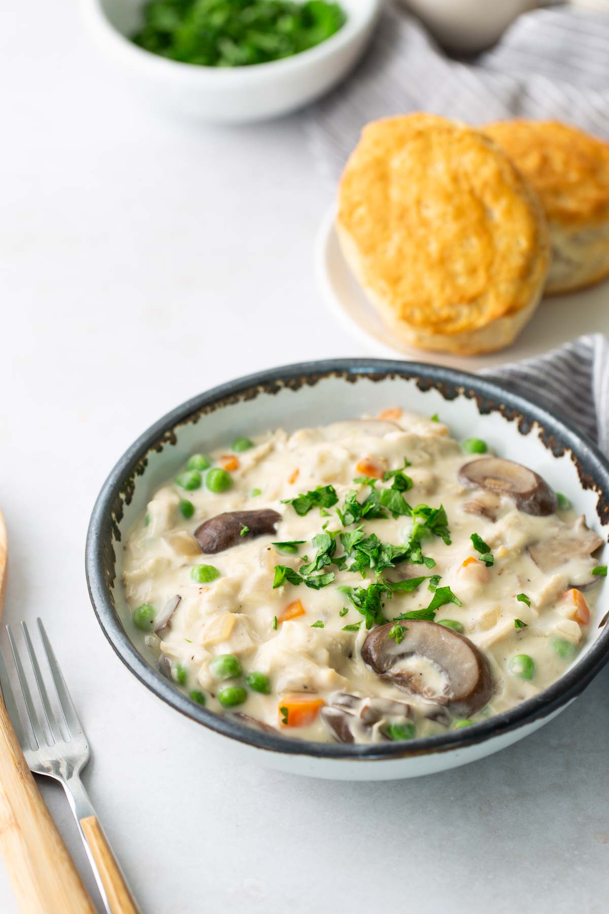 A bowl of creamy vegetable and chicken soup topped with parsley, next to a plate with two biscuits.