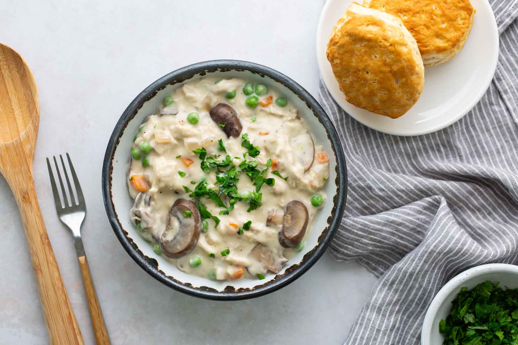 A bowl of creamy chicken stew with peas, carrots, and mushrooms, garnished with parsley, next to two biscuits on a plate. A wooden spoon and fork are placed nearby.