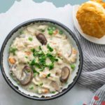 A bowl of creamy turkey à la king with mushrooms, peas, and carrots, garnished with parsley. Biscuits are on the side, placed on a striped cloth.