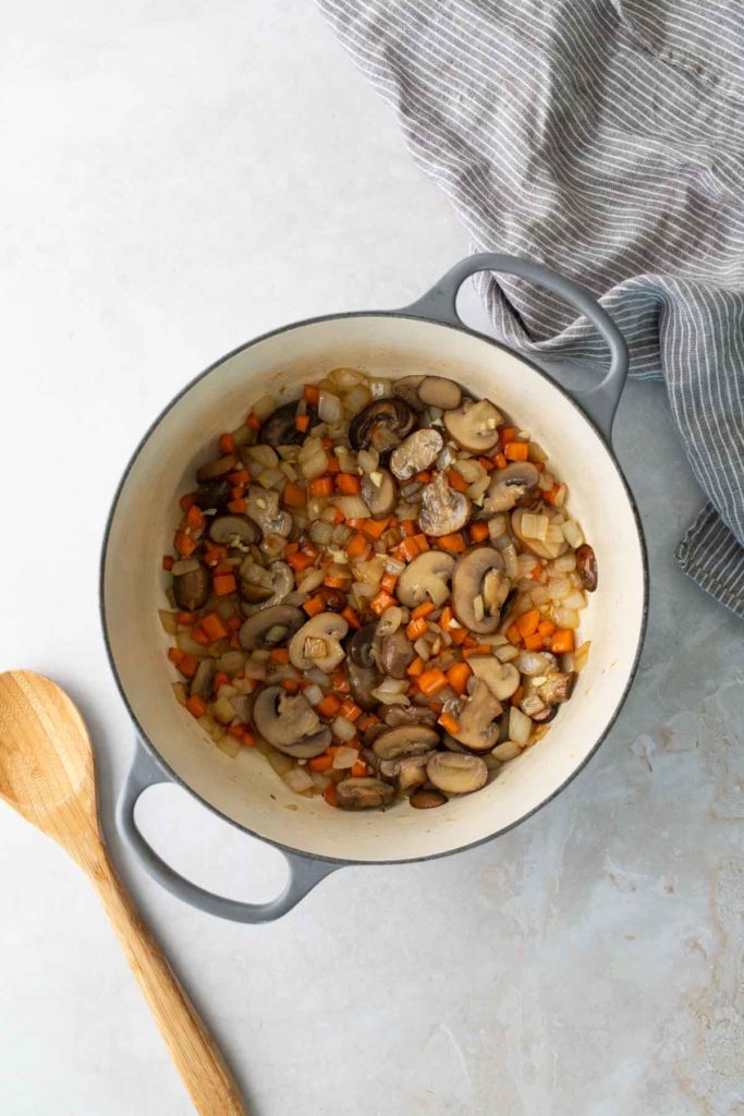 A pot of sautéed mushrooms, onions, and carrots on a light surface, with a wooden spoon and striped cloth nearby.