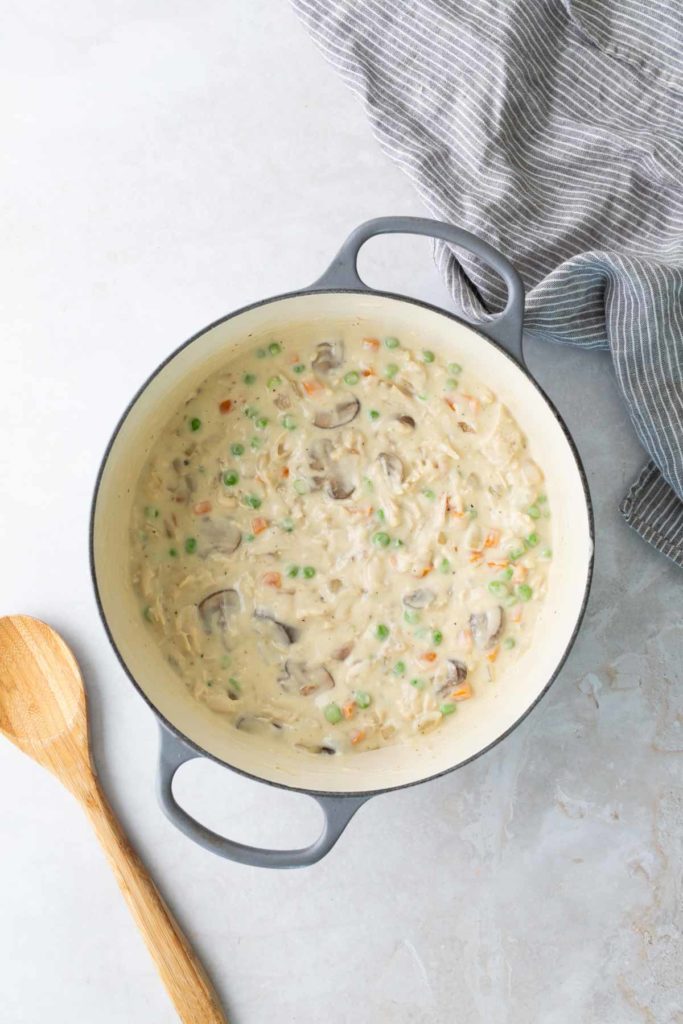 A pot of creamy mushroom soup with peas and carrots, next to a wooden spoon on a light surface.