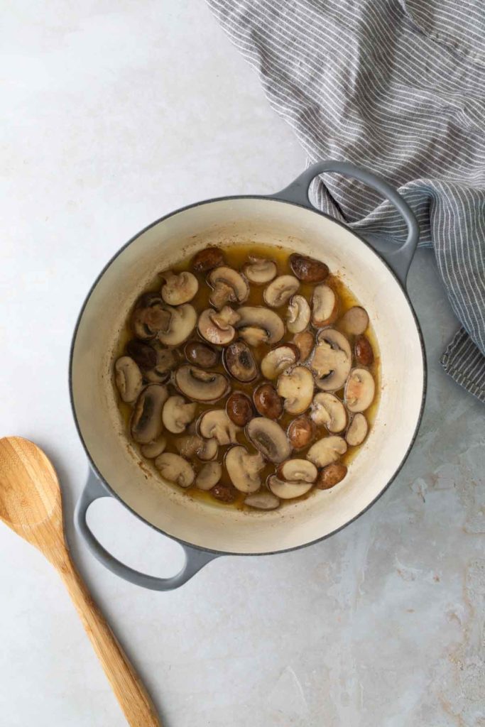 Sliced mushrooms cooking in a light-colored pot on a gray surface, with a wooden spoon and striped cloth nearby.