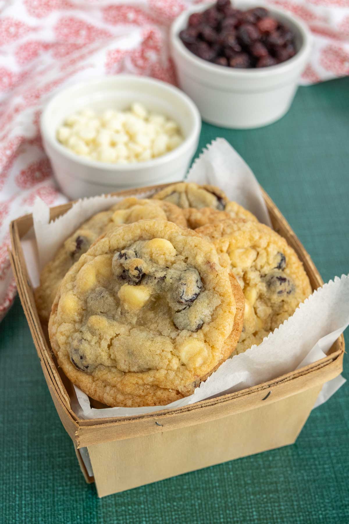 A basket with white chocolate chip and cranberry cookies, accompanied by small bowls of white chocolate chips and dried cranberries in the background.