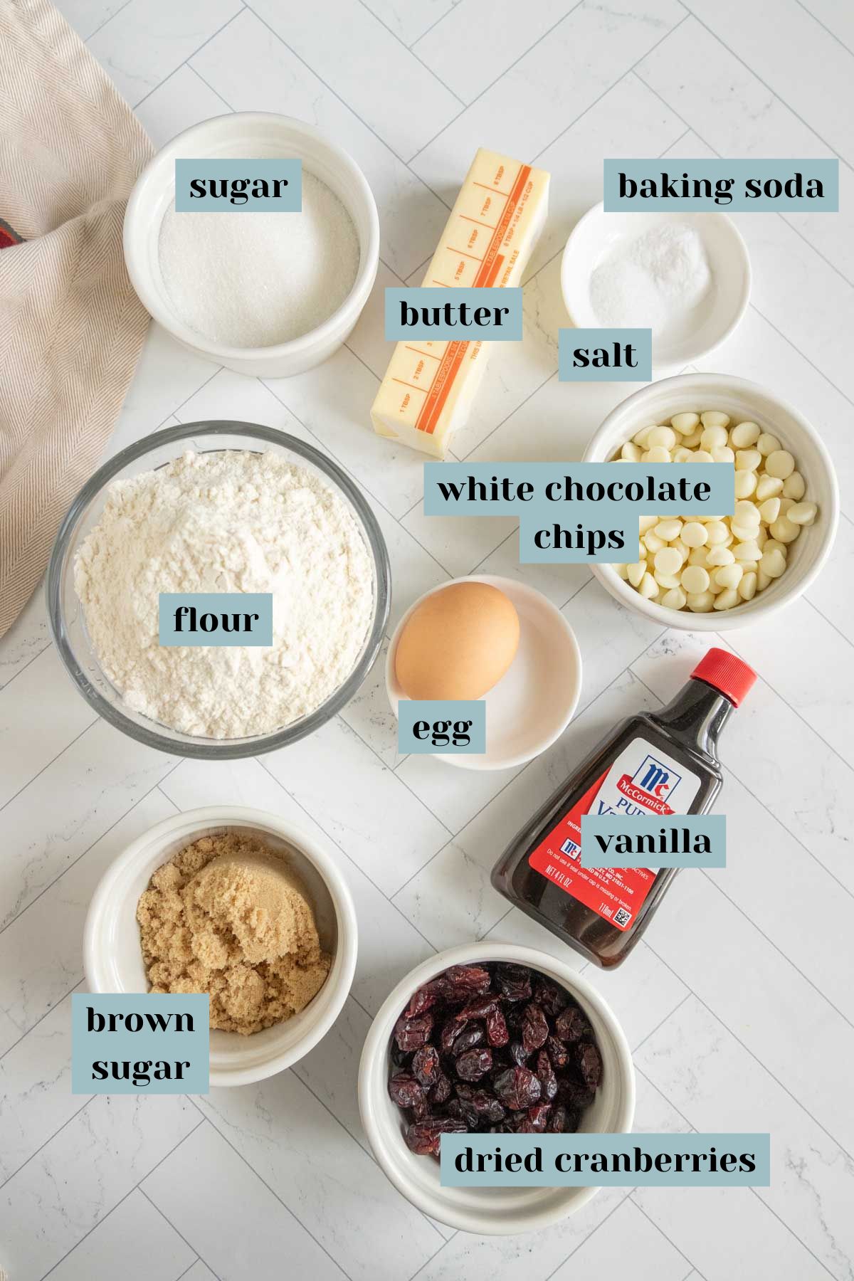 Ingredients for baking are arranged on a white surface, including sugar, butter, baking soda, salt, white chocolate chips, flour, egg, vanilla, brown sugar, and dried cranberries.