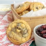 A wicker basket of cookies with chocolate chips on a red-patterned napkin, a cookie leaning on it. Nearby, a small bowl of dried fruit and another of white chocolate chips.