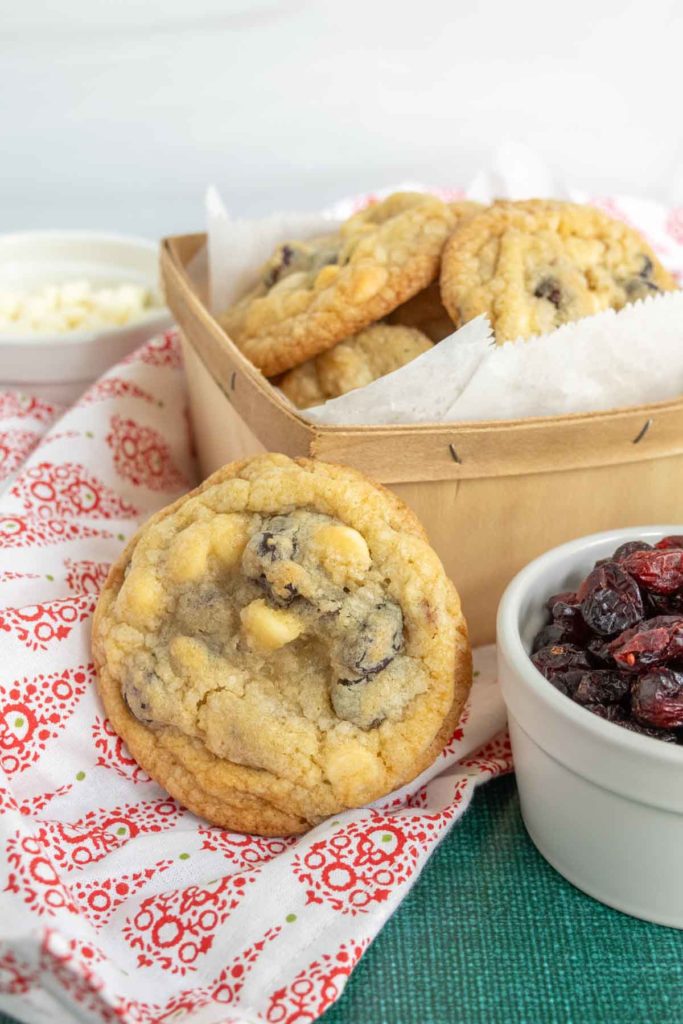 A wicker basket of cookies with chocolate chips on a red-patterned napkin, a cookie leaning on it. Nearby, a small bowl of dried fruit and another of white chocolate chips.