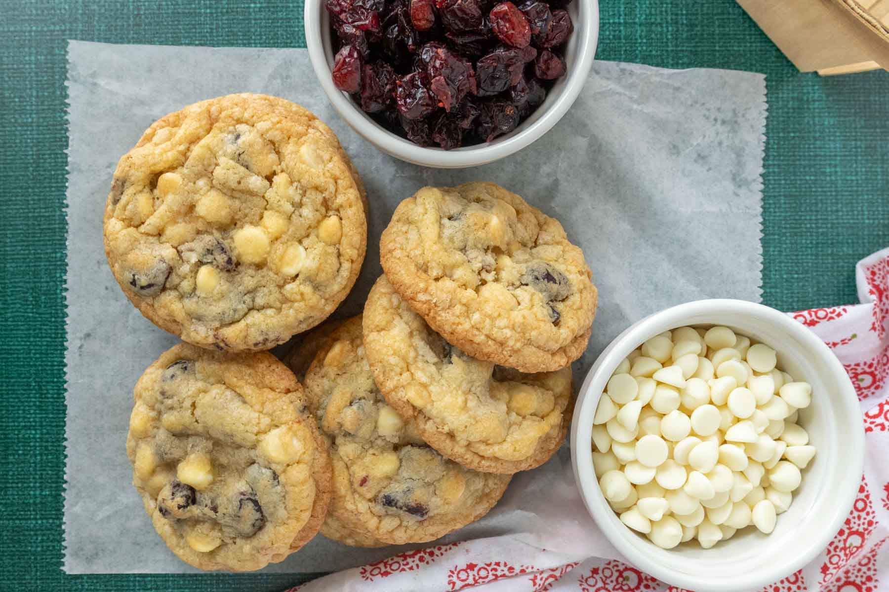 A stack of cookies with white chocolate chips on parchment paper, next to bowls of dried cranberries and white chocolate chips.