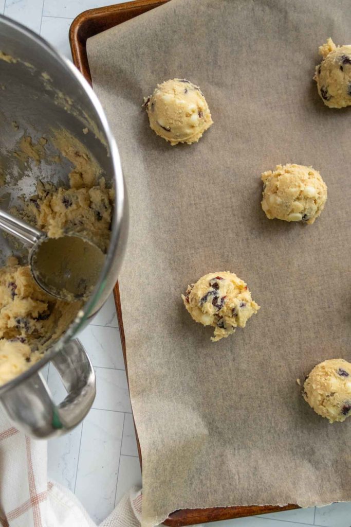 Cookie dough portions on a parchment-lined baking sheet next to a mixing bowl with a scoop.