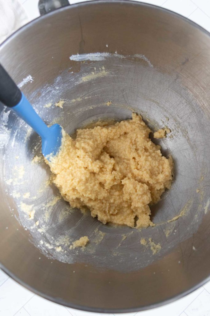 A blue spatula in a metal bowl containing a yellow, crumbly dough mixture.