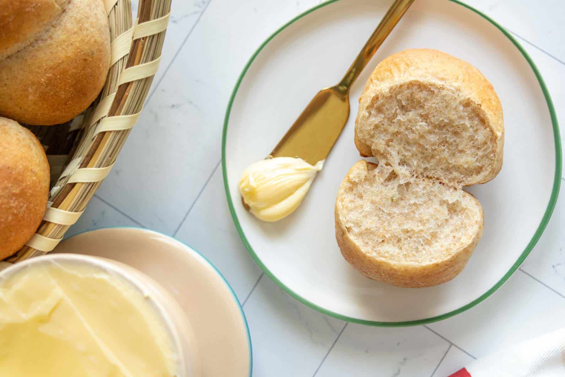 A sliced bread roll on a plate with a small butter portion and a butter knife, next to a basket of rolls and a butter dish.