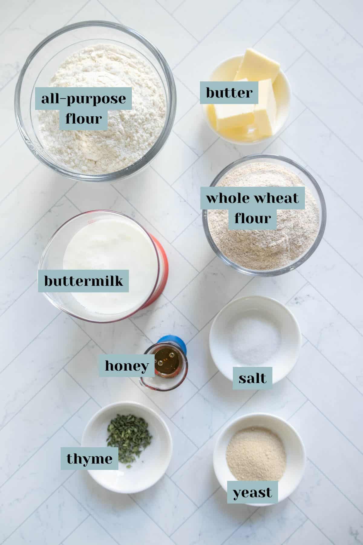 Ingredients for breadmaking: all-purpose flour, whole wheat flour, butter, buttermilk, honey, salt, thyme, and yeast, arranged on a white surface.