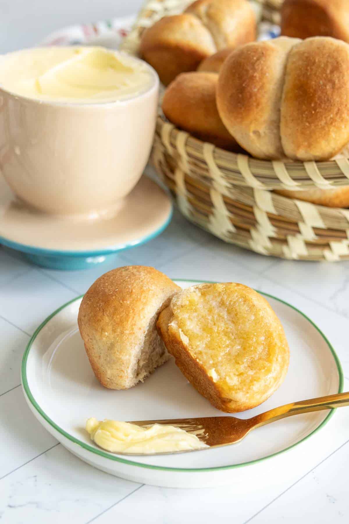 A plate with a partially buttered roll, a knife with butter, and a basket of rolls in the background. A cup of butter is beside the basket.