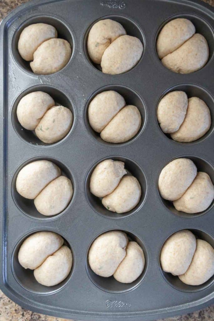 A muffin tin with six compartments, each holding unbaked dough shaped into rolls, ready for baking.