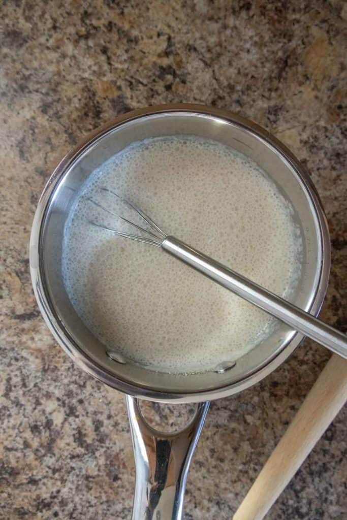 A small saucepan filled with a creamy liquid, a whisk resting inside. It's placed on a brown granite countertop.