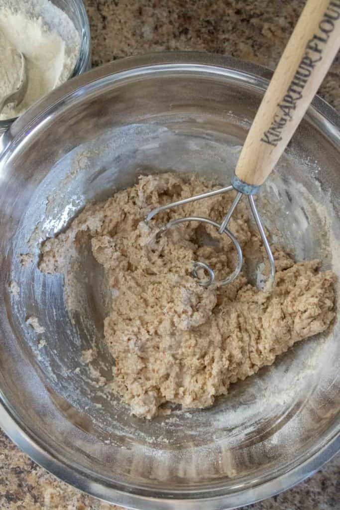 A metal bowl with dough being mixed using a wooden-handled utensil on a speckled countertop. A bowl of flour is visible in the background.