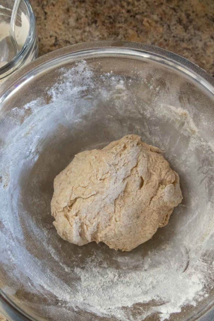 A ball of dough in a floured glass bowl on a brown countertop.