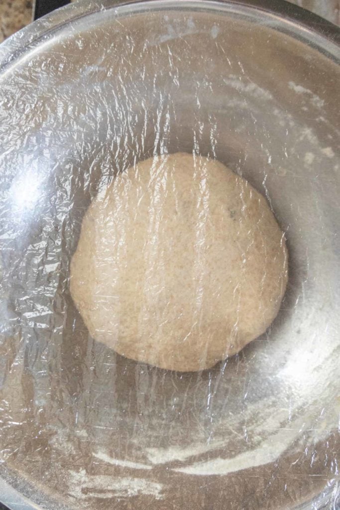 A ball of dough covered with plastic wrap in a stainless steel bowl.