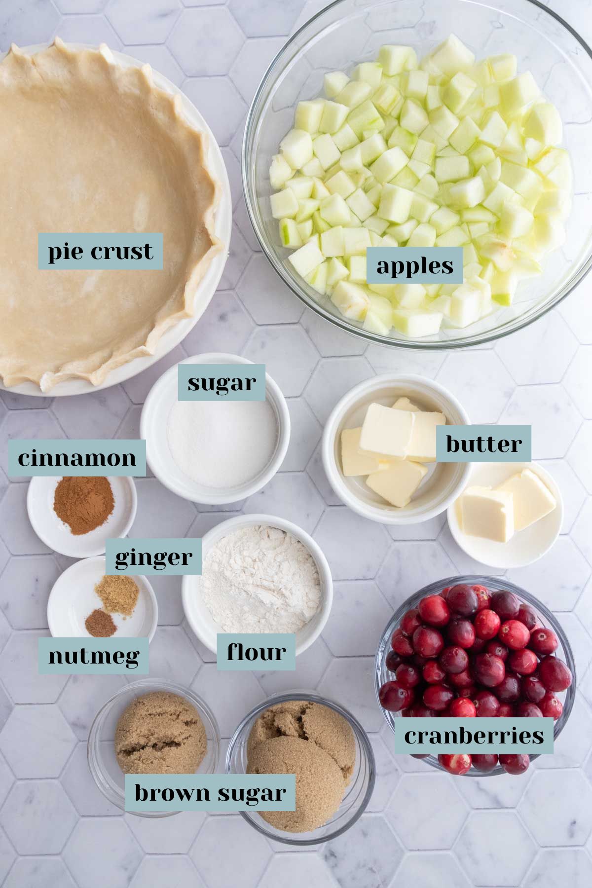 Overhead view of labeled pie ingredients on a marble surface, including pie crust, apples, sugar, butter, cranberries, brown sugar, flour, cinnamon, ginger, and nutmeg.