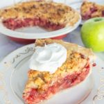 A slice of crumble-topped fruit pie with whipped cream on a decorative plate, next to a whole pie with a slice missing, and a green apple on the side.