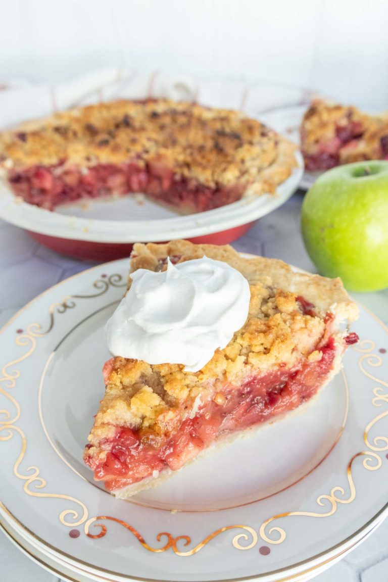 A slice of crumble-topped fruit pie with whipped cream on a decorative plate, next to a whole pie with a slice missing, and a green apple on the side.